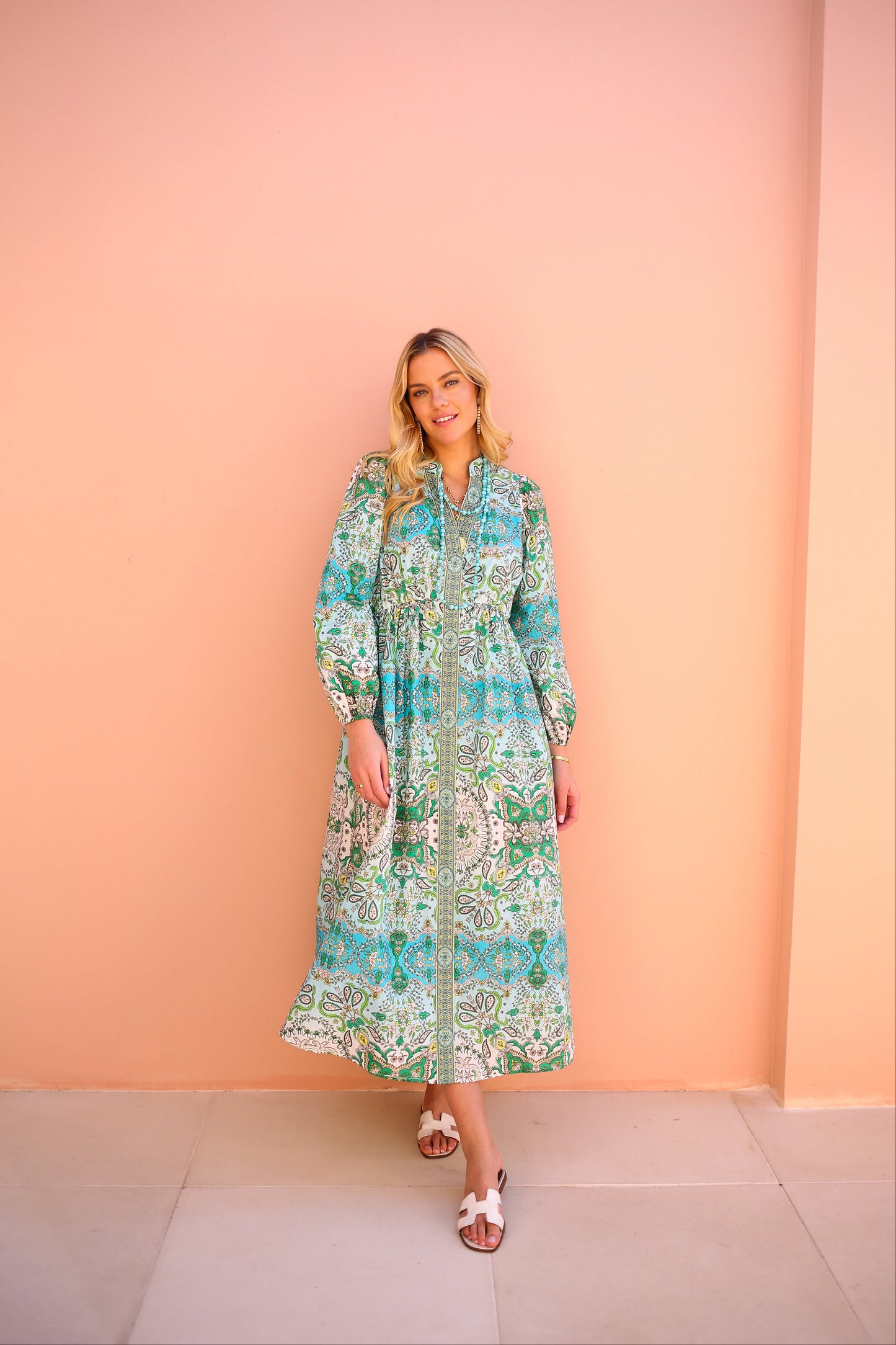 Woman wearing a long, patterned dress against a peach-colored wall.