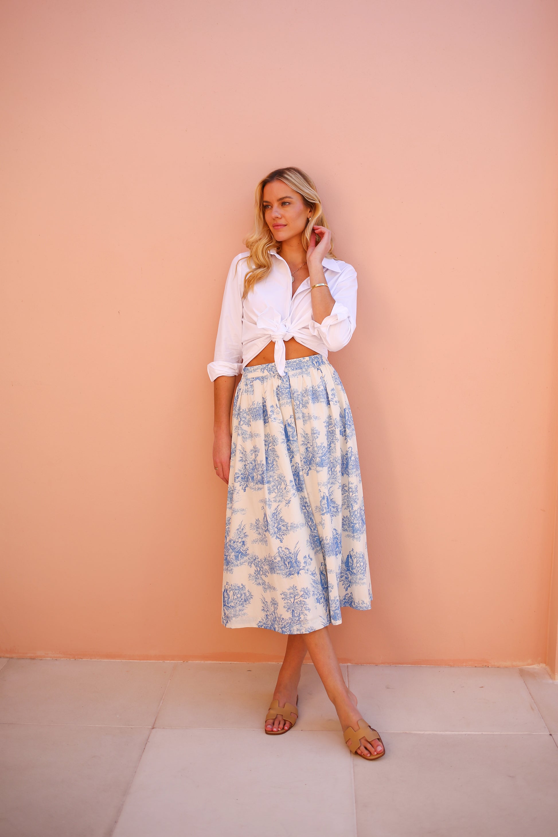 Woman wearing a white blouse and floral skirt against a peach wall.
